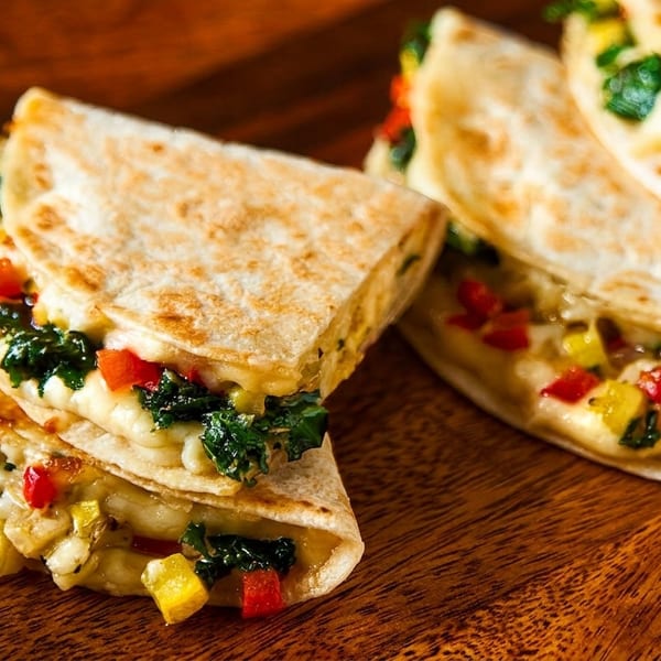 Cheesy quesadillas filled with colorful vegetables, including red and yellow peppers and green spinach, on a wooden board.