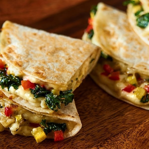 Golden-brown quesadillas filled with melted cheese, green kale, and diced red and yellow bell peppers on a wooden board.