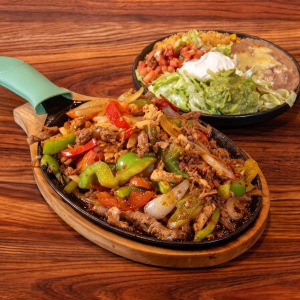 Sizzling fajitas with bell peppers and onions, alongside a plate of rice, beans, guacamole, and sour cream.