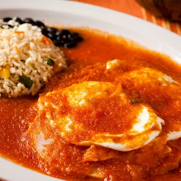 Huevos rancheros with two fried eggs in red sauce, rice, and black beans on a white plate.