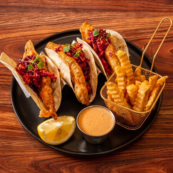 Three fish tacos with red cabbage slaw, a side of crinkle-cut fries, sauce, and lemon.